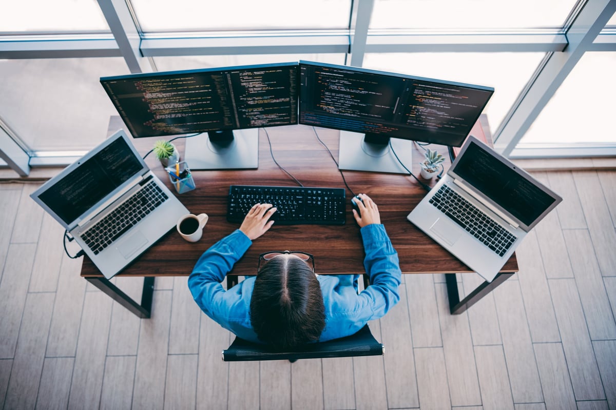 A focused programmer works at a spacious desk with three monitors and two laptops in a bright loft office, showcasing coding concentration and collaborative tech work.